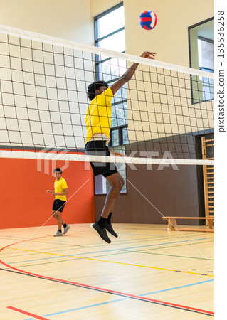 Diverse male teammates jumping next to net intercepting red white blue volleyball in gym 135536258
