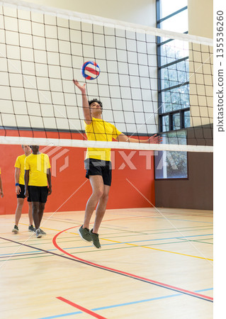 Diverse male teammates jumping, reaching volleyball over net in yellow jerseys on court Diverse male teammates jumping, reaching volleyball over net in yellow jerseys on court 135536260
