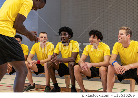 Diverse male teammates talking around wooden bench in gymnasium with court lines, yellow jerseys 135536269