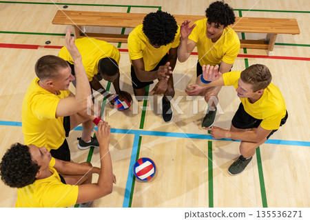 Diverse male teammates huddling around volleyball on gym court wearing yellow tees, black shorts 135536271