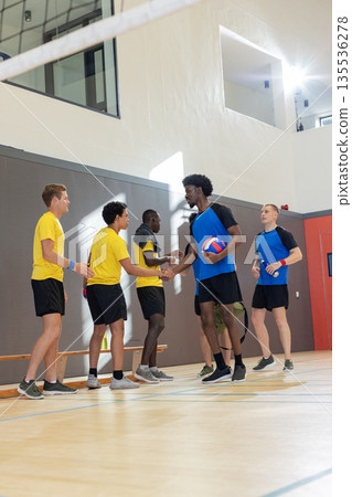 Diverse male athletes in jerseys shaking hands at center court holding volleyball near net Diverse male athletes in jerseys shaking hands at center court holding volleyball near net 135536278