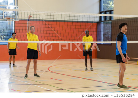 Diverse male volleyball athletes jumping and blocking on gym court with net, lines 135536284