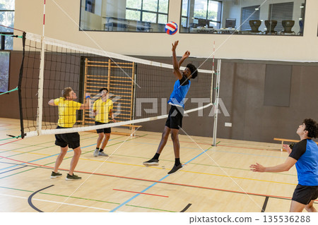 Diverse male players spiking volleyball over net in yellow and blue jerseys in gym, copy space 135536288