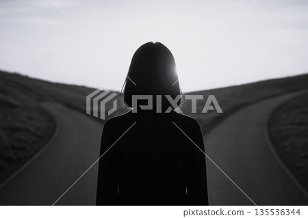 A black-and-white photograph of a woman standing on a hill at dusk, facing a crossroads in her life and a fork in the road. 135536344