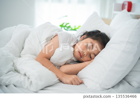 Young girl peacefully sleeping on a cozy bed surrounded by soft pillows in a bright and airy room, emphasizing calm and comfort in a serene setting. 135536466
