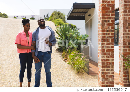 Couple standing outside modern house, smiling and enjoying sunny day together Couple standing outside modern house, smiling and enjoying sunny day together 135536947
