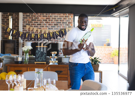Man celebrating retirement holding wine bottle and smiling in decorated living room Man celebrating retirement holding wine bottle and smiling in decorated living room 135536977