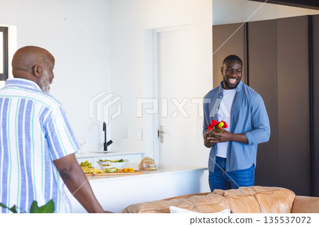 Man holding flowers smiling at older man in modern kitchen, celebrating together 135537072