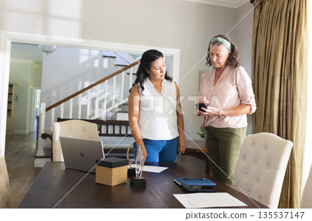 Two women discussing smartphone features at home office with laptop and documents 135537147