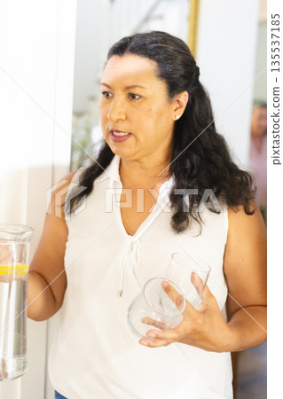 Woman holding water pitcher and glass, engaging in conversation at home gathering Woman holding water pitcher and glass, engaging in conversation at home gathering 135537185