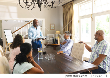 Senior man presenting on tablet to group at home, everyone smiling and engaged 135537191