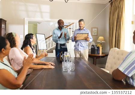 Senior man receiving award at home, surrounded by friends clapping and celebrating 135537212