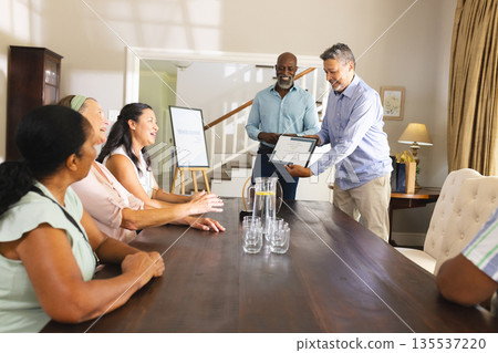 Receiving certificate at home, senior man surrounded by applauding friends at table 135537220