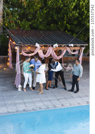 Exchanging hugs and flowers, diverse group celebrating by poolside under pergola 135537342