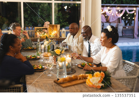Seniors enjoying dinner party, sharing laughter and wine by poolside at night 135537351