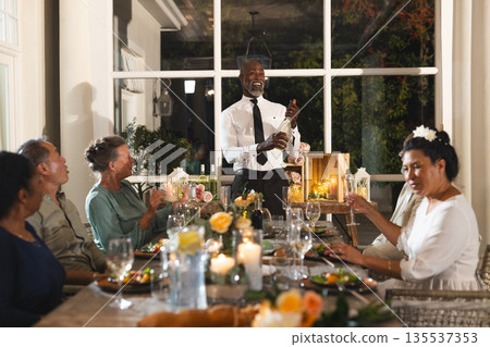Man opening champagne bottle at elegant dinner party, guests smiling and celebrating 135537353