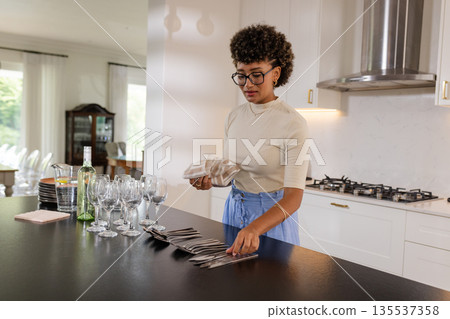African American woman arranging glassware and cutlery in modern kitchen for event 135537358