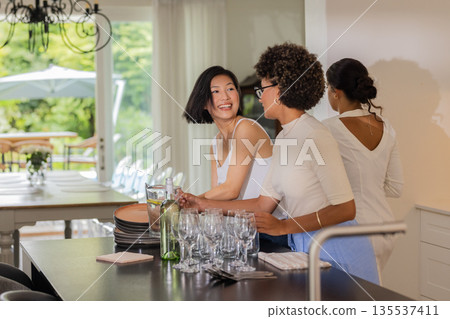 Women preparing tableware and glasses, smiling and chatting in bright dining area Women preparing tableware and glasses, smiling and chatting in bright dining area 135537411