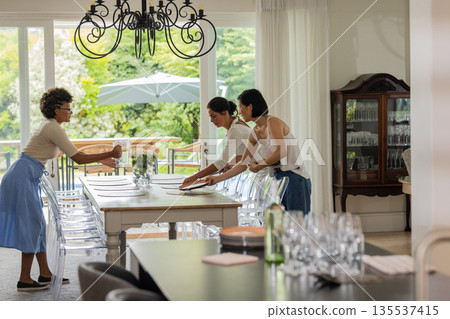 Women setting elegant dining table for wedding celebration in bright, airy room Women setting elegant dining table for wedding celebration in bright, airy room 135537415