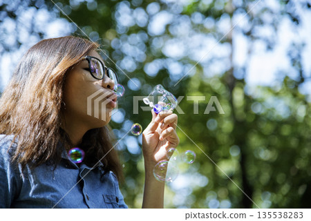 Adorable happy cute girl playful foam bubbles in green playground in summer outdoors. Funny Cheerful girl in the park happiness times. Bubbles blowing soap playful in nature park. 135538283