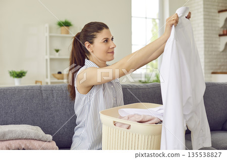Joyful woman sitting at home on sofa and holding clean laundry clothes in basket Joyful woman sitting at home on sofa and holding clean laundry clothes in basket 135538827