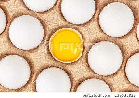 Cracked egg with bright yolk among white eggs in cardboard tray top view with graphic pattern Cracked egg with bright yolk among white eggs in cardboard tray top view with graphic pattern 135539825