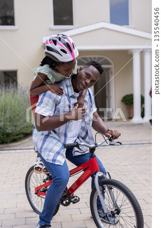 Father riding bicycle with daughter wearing helmet, enjoying playful outdoor moment Father riding bicycle with daughter wearing helmet, enjoying playful outdoor moment 135540456