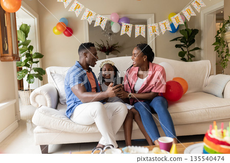 Celebrating birthday at home, African American family smiling and looking at smartphone Celebrating birthday at home, African American family smiling and looking at smartphone 135540474