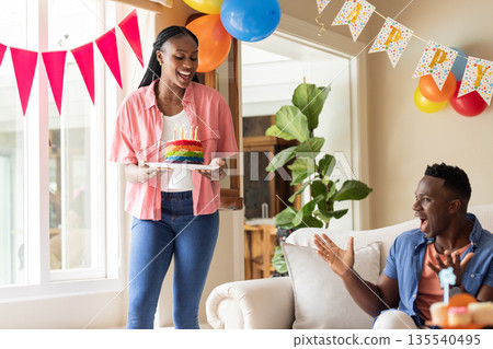 African American woman bringing birthday cake to surprised man at home celebration 135540495