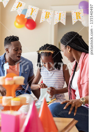 Family celebrating birthday at home, child opening gift with parents watching Family celebrating birthday at home, child opening gift with parents watching 135540497