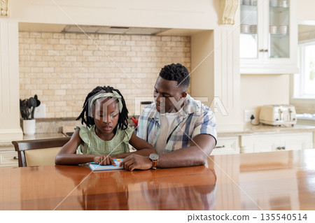 Father and daughter reading tablet together at kitchen table, sharing quality time Father and daughter reading tablet together at kitchen table, sharing quality time 135540514
