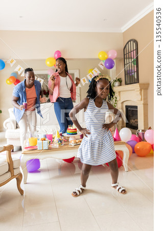 Family celebrating birthday at home, young girl dancing with parents cheering Family celebrating birthday at home, young girl dancing with parents cheering 135540536