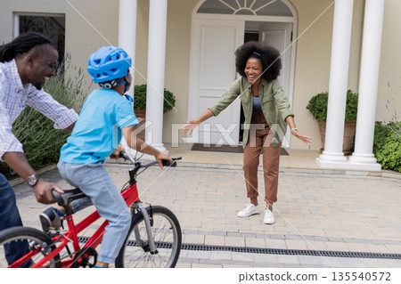 Father guiding child on bicycle towards smiling mother outside home Father guiding child on bicycle towards smiling mother outside home 135540572