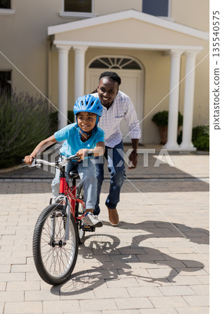 African American father teaching son to ride bicycle outside, both smiling happily 135540575