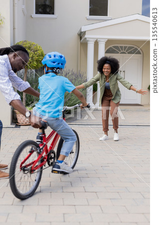 Child riding bicycle towards parents, smiling and encouraging outside family home Child riding bicycle towards parents, smiling and encouraging outside family home 135540613