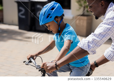 African American father teaching son to ride bicycle outdoors, both enjoying moment 135540629
