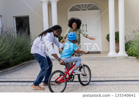 African American parents teaching child to ride bicycle outside home, all smiling 135540641