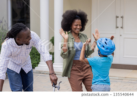 Child wearing helmet high-fiving mom, African American family playing outdoors 135540644