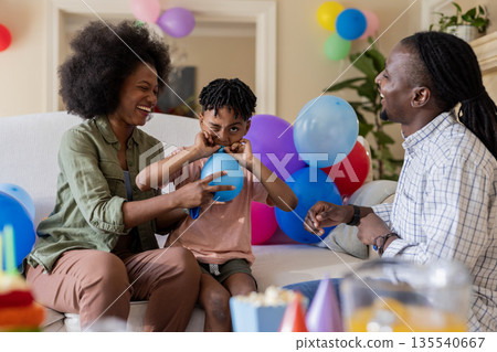 Family celebrating birthday at home, child blowing balloon with parents smiling Family celebrating birthday at home, child blowing balloon with parents smiling 135540667