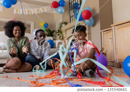 Family celebrating birthday at home, child playing with colorful streamers on floor Family celebrating birthday at home, child playing with colorful streamers on floor 135540681