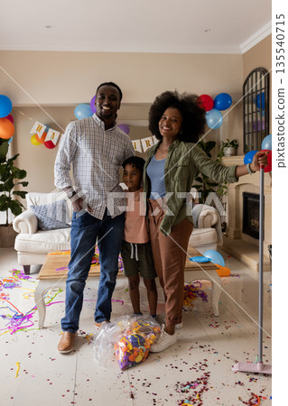 Family celebrating birthday at home, smiling with balloons and confetti on floor Family celebrating birthday at home, smiling with balloons and confetti on floor 135540715