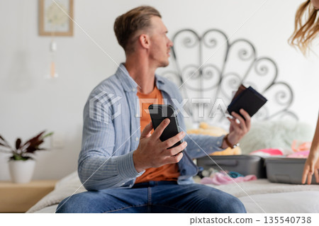 Man packing suitcase holding smartphone and wallet, preparing for travel at home 135540738