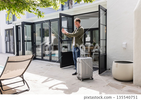 Man taking selfie with smartphone by poolside, standing with suitcase outdoors 135540751