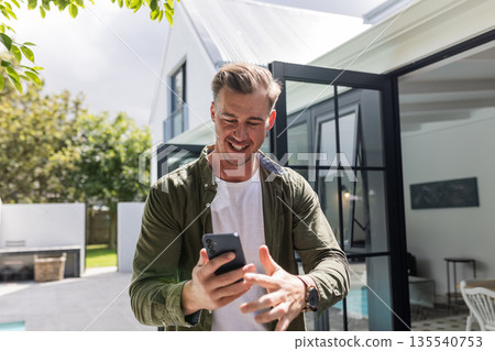 Smiling man using smartphone outside modern home, enjoying sunny day Smiling man using smartphone outside modern home, enjoying sunny day 135540753