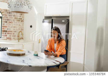 At home, woman using smartphone in modern kitchen, sitting at marble counter 135540784