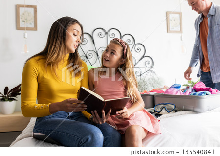 Mother and daughter reading book together on bed while father packs suitcase Mother and daughter reading book together on bed while father packs suitcase 135540841