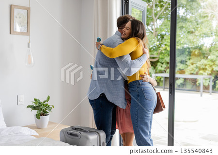 Family embracing warmly by open patio door, suitcase on floor, enjoying reunion Family embracing warmly by open patio door, suitcase on floor, enjoying reunion 135540843