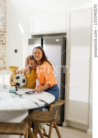 Mother and daughter hugging in kitchen with soccer ball and trophy Mother and daughter hugging in kitchen with soccer ball and trophy 135540870