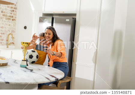 Mother and daughter taking selfie with soccer trophy in kitchen, smiling happily Mother and daughter taking selfie with soccer trophy in kitchen, smiling happily 135540871
