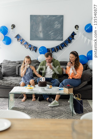 Family celebrating birthday at home, enjoying cake and drinks on sofa 135540877